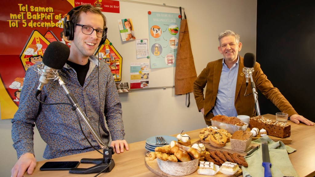 Zegert staat links met een kleurrijke bloes en een koptelefoon op. Frans staat rechts, met een bruin jasje en een blauw overhemd. Ze lachen. Voor hen staat een tafel met allerlei brood en gebak. Ze hebben allebei een grote microfoon voor zich.